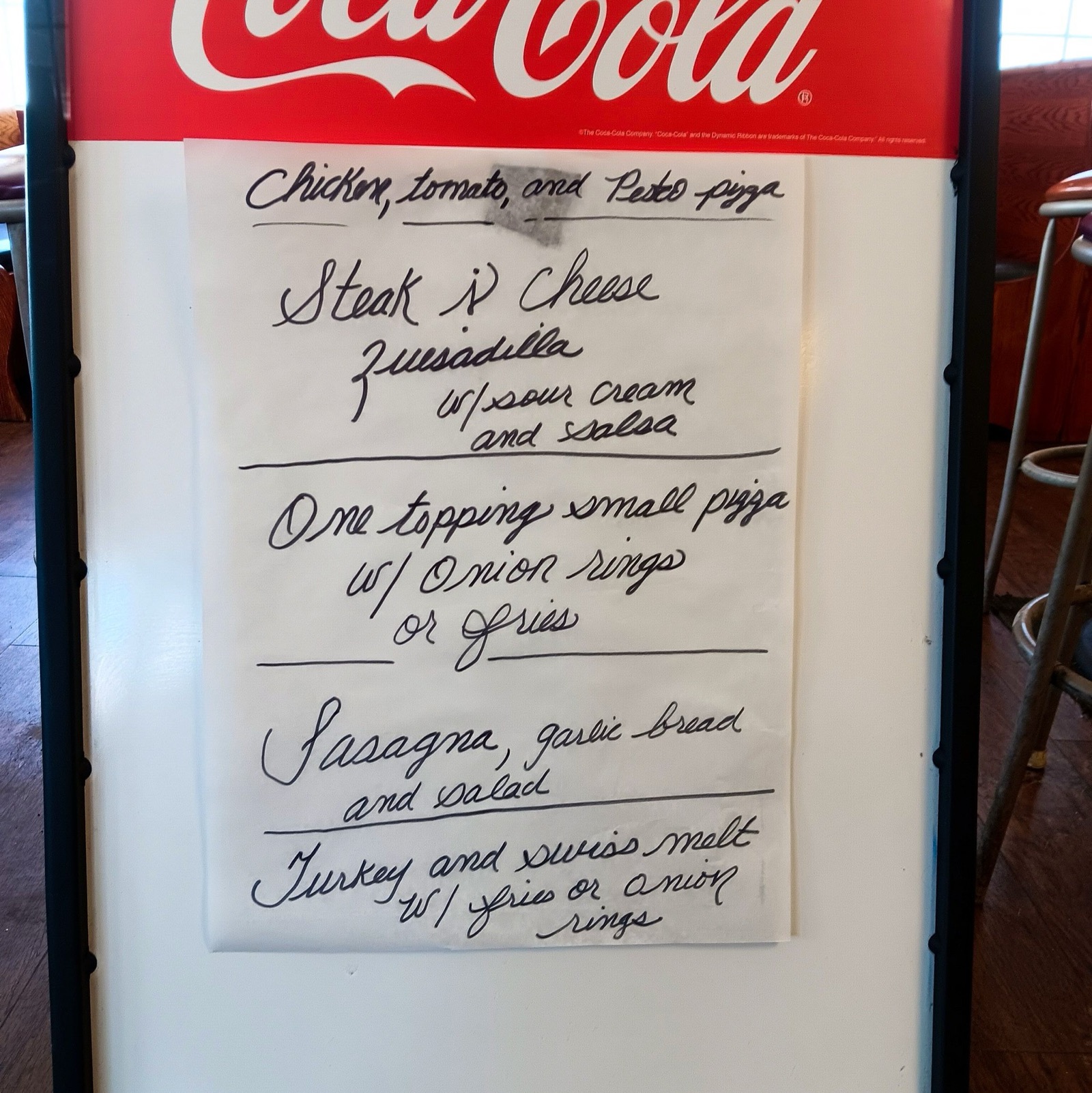 A Coca-Cola whiteboard with handwritten specials: chicken, tomato and pesto pizza, steak and cheese quesadilla, one topping small pizza with onion rings or fries, lasagna with garlic bread and salad, turkey and Swiss melt