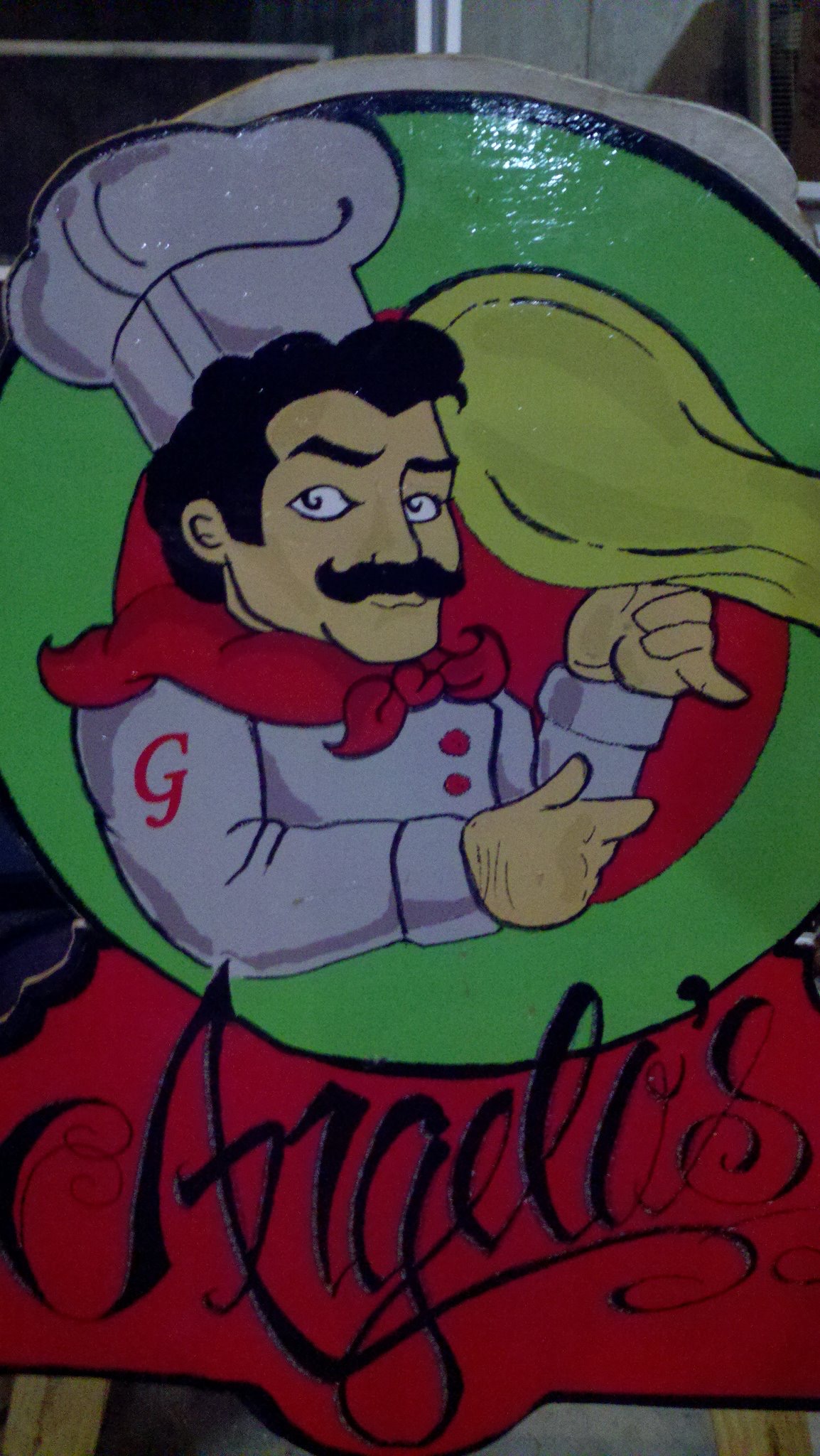 The shop’s hand-painted sign: a mustached chef with a G on his coat tossing pizza dough, above the name Angelo’s in black script on red