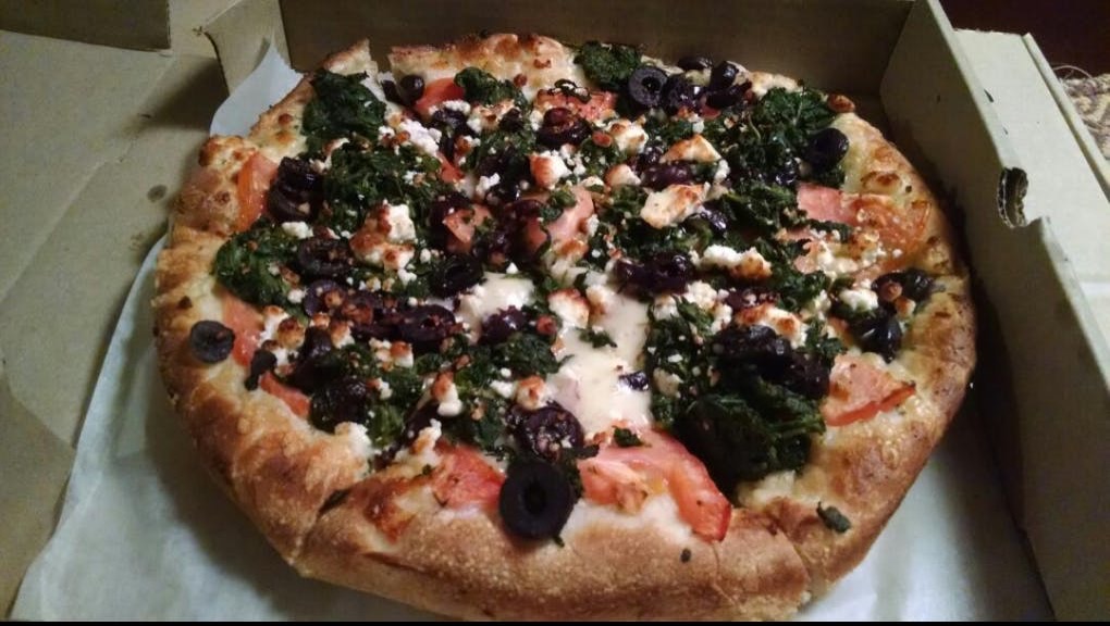A Greek pizza in its box, topped with spinach, black olives, tomato and feta