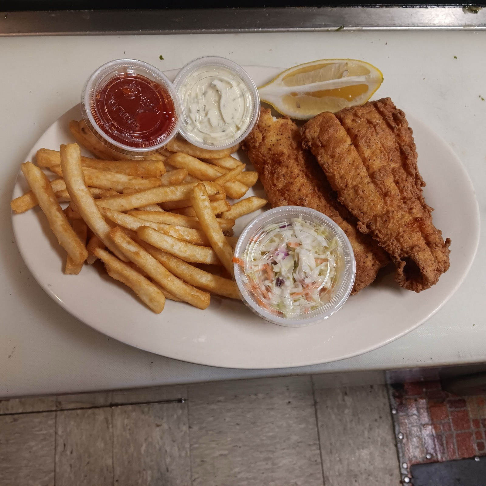 Fried haddock with French fries, coleslaw, tartar sauce, ketchup and a lemon wedge on a white oval plate