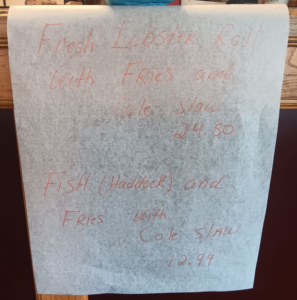 A handwritten sign offering a fresh lobster roll with fries and coleslaw for 24.50 and fish (haddock) and fries with coleslaw for 12.99