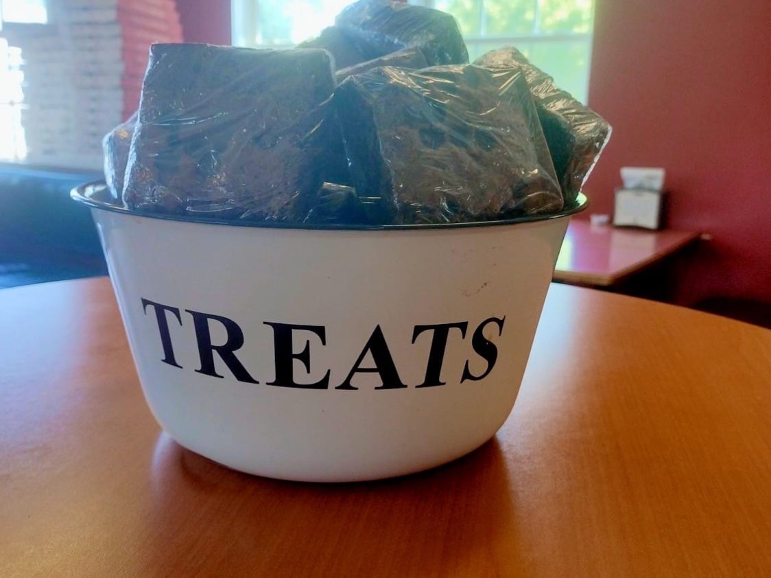 A white enamel bowl labeled TREATS, full of wrapped brownies, on a table by the window